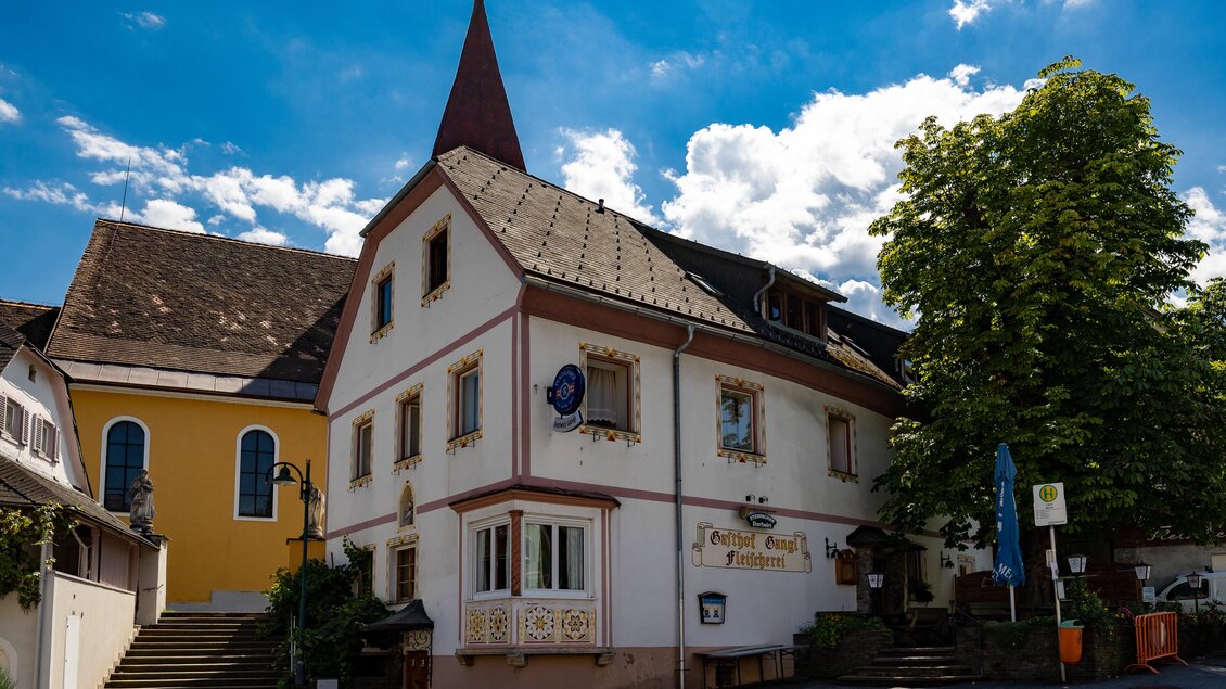 Außenansicht des Gasthauses Gangl in Ligist mit markantem Giebel, Gastgarten und angrenzender Kirche. | © TV Region Graz - Harry Schiffer