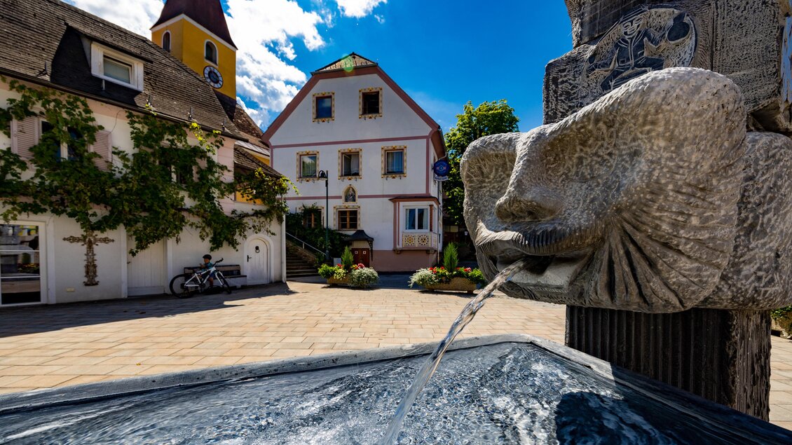 Brunnen mit steinernem Wasserspeier vor dem Gasthaus Gangl und der Kirche im Ortszentrum von Ligist. | © TV Region Graz - Harry Schiffer