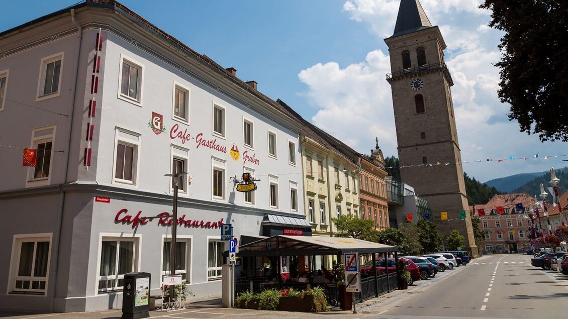 Eine charmante Straßenszene mit einem Café-Restaurant und einem hohen Kirchturm. Der Himmel ist blau mit einigen Wolken und die Gebäude sind gut erhalten. | © Gasthaus Gruber Hubmann