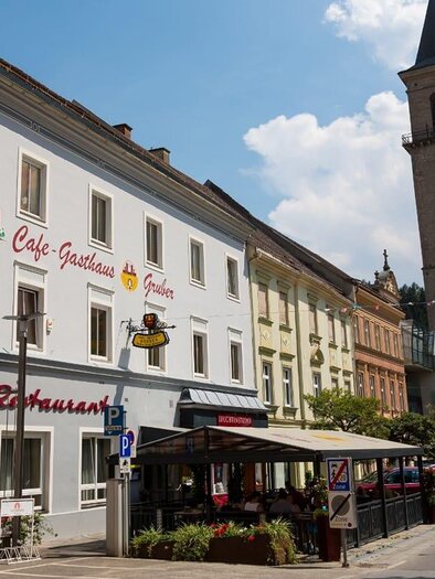 Eine charmante Straßenszene mit einem Café-Restaurant und einem hohen Kirchturm. Der Himmel ist blau mit einigen Wolken und die Gebäude sind gut erhalten. | © Gasthaus Gruber Hubmann