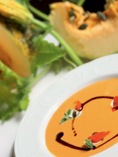 Eine köstliche Kürbiscremesuppe in einer weißen Schüssel. Im Hintergrund liegt ein aufgeschnittener Kürbis. | © Graz Tourismus-Harry Schiffer