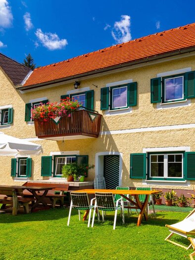A charming house with green shutters in a sunny setting. In the foreground, there are tables, chairs, and loungers on a green lawn. | © TV Region Graz - Harry Schiffer
