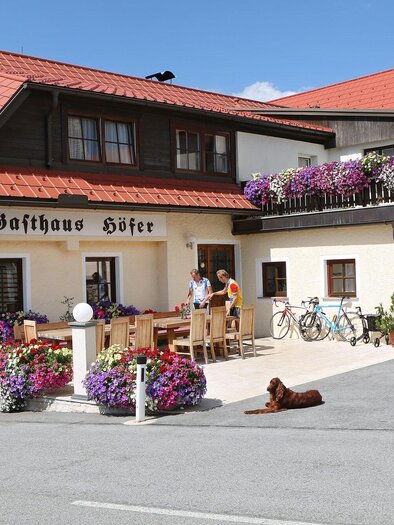 An inviting inn with colorful flower boxes and a cozy terrace. Bicycles are ready, and a dog is lounging relaxed in front. | © TV Region Graz - Donner
