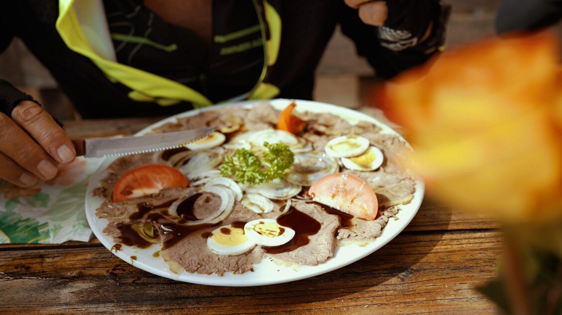Ein Teller mit Carpaccio, garniert mit Zwiebeln, Eiern und frischen Tomaten. Im Hintergrund ist eine Blume zu sehen. | © TV Region Graz - Miasphotoart