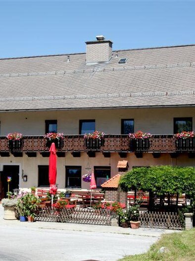 Ein gemütliches Gasthaus mit Balkon und blühenden Pflanzkübeln. Es liegt umgeben von Bäumen und einer schönen Landschaft.