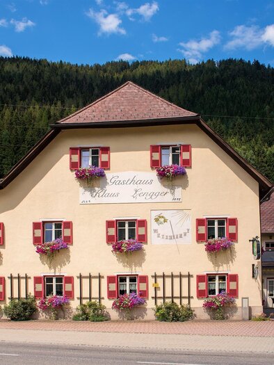 Ein charmantes Gasthaus mit bunten Fensterläden und vielen Blumen. Im Hintergrund sind grüne Hügel und ein blauer Himmel zu sehen. | © FVV Tragöß St. Katharein