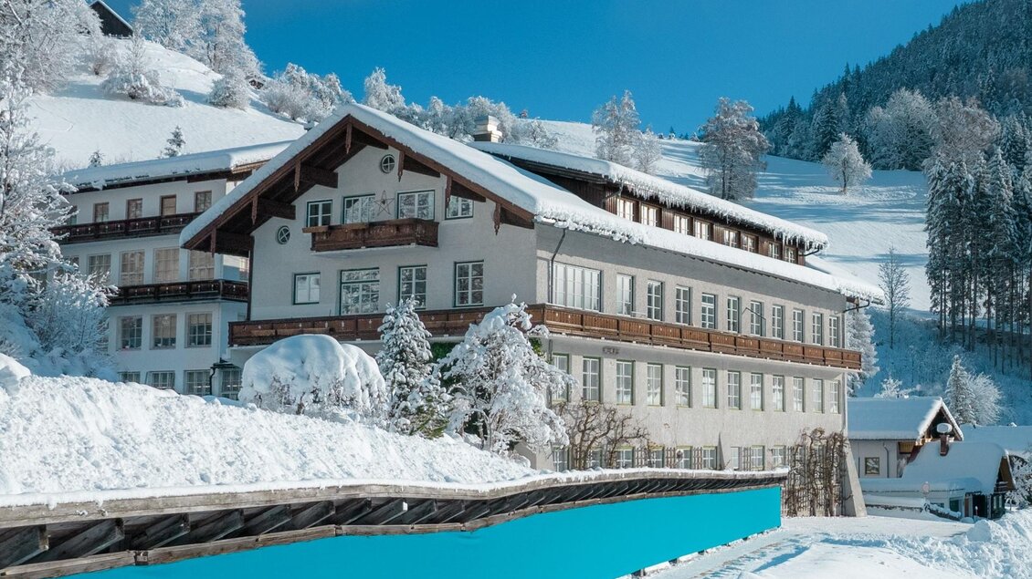Außenansicht im Winter vom Lodenwalker Ramsau, bei strahlend blauem Himmel und schneebedeckter Landschaft. Vor dem Gebäude ist eine lange Stoffbahn aus blauem Loden aufgehängt um ihn an der frischen Luft zu trocknen. | © Lodenwalker Ramsau
