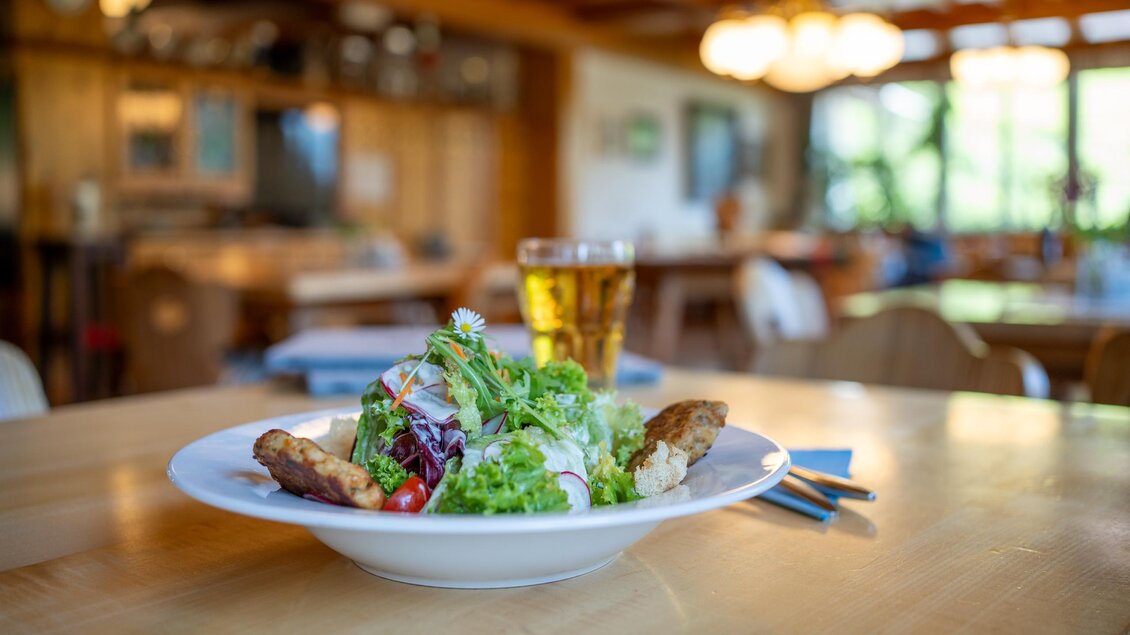Kaspressknödel auf knackigen Blattsalaten. Im Hintergrund ist ein Glas Apfelsaft und eine gemütliche Restaurantatmosphäre zu sehen. | © Lodenwalker Ramsau