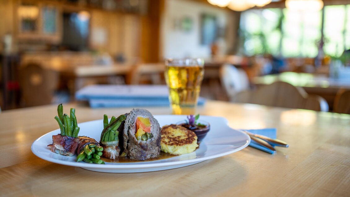 Zart gedünstete Rindsroulade mit Rotweinsauce, Erdäpfelkrapferl & Speckbohnen. Im Hintergrund ist ein Glas Apfelsaft und eine gemütliche Restaurantatmosphäre zu sehen. | © Lodenwalker Ramsau