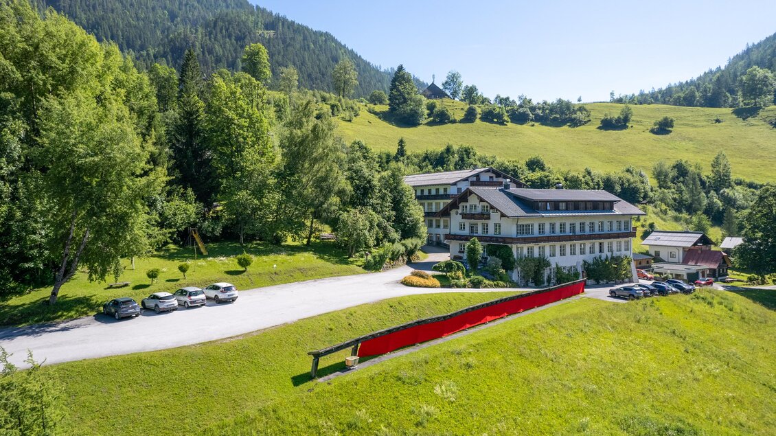 Außenansicht vom Lodenwalker Ramsau im Sommer. Parkplatz und Spielplatz sind gut sichtbar. Im Vordergrund ist eine lange Stoffbahn aus rotem Loden zu sehen, der an der frischen Luft trocknet. | © Lodenwalker Ramsau