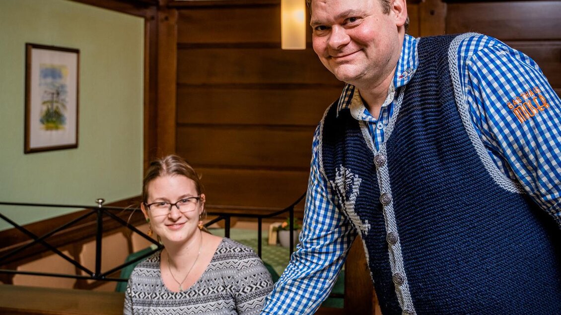 Ein Restaurantbesuch mit einem fröhlichen Kellner, der ein Gericht serviert. Im Hintergrund sitzt eine zufriedene Frau am Tisch. | © Stephan Friesinger