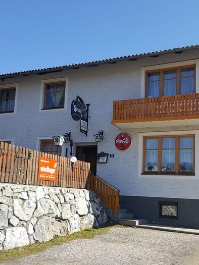 Ein Gasthaus mit einem Erdgeschoss und einem Balkon. Im Hintergrund sind Berge und ein blauer Himmel sichtbar. | © Gasthaus Mossauer