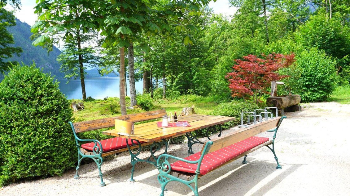 Ein einladender Platz mit einer Holztafel und Bänken, umgeben von Bäumen und bunten Pflanzen. Im Hintergrund ist ein ruhiger See zu sehen. | © Peter Prommer