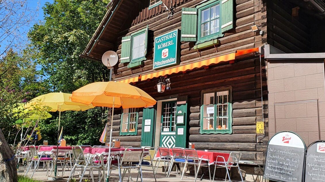 Ein gemütliches Gasthaus mit Holzfassade und grünen Fensterläden. Tische mit bunten Sonnenschirmen stehen im Freien.