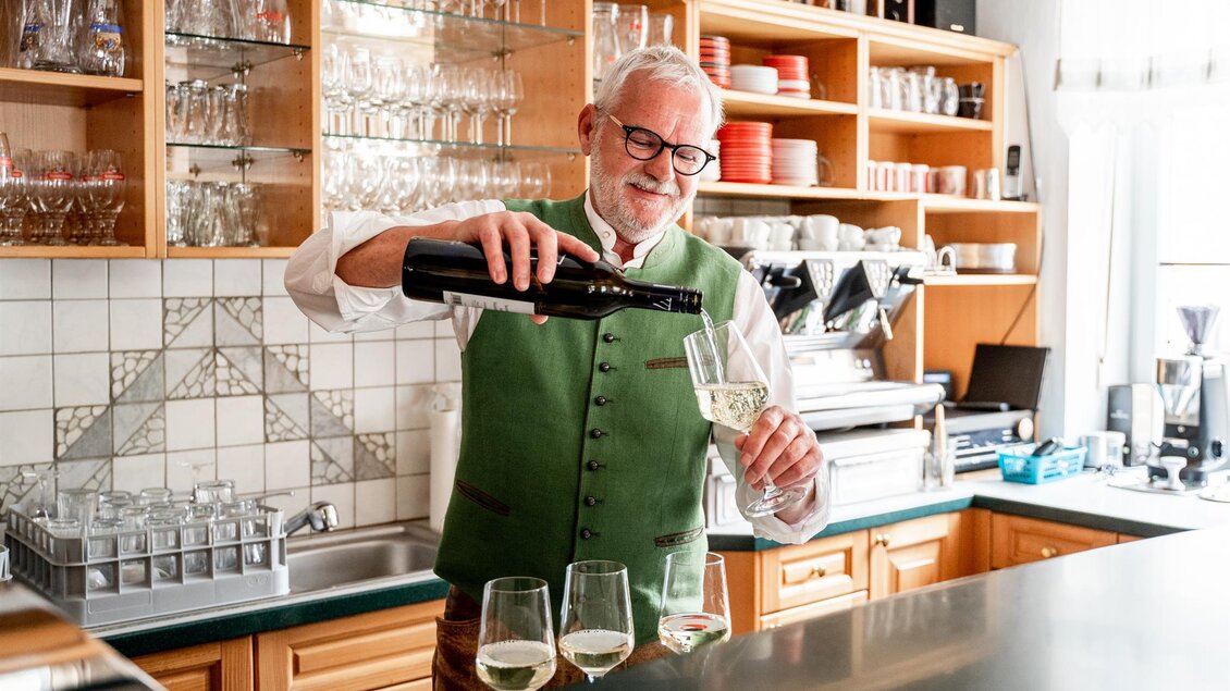 Gastgeber in grüner Weste schenkt Weißwein hinter der Theke im Platzwirt in Graden ein. | © Platzwirt_KerstinFotografie
