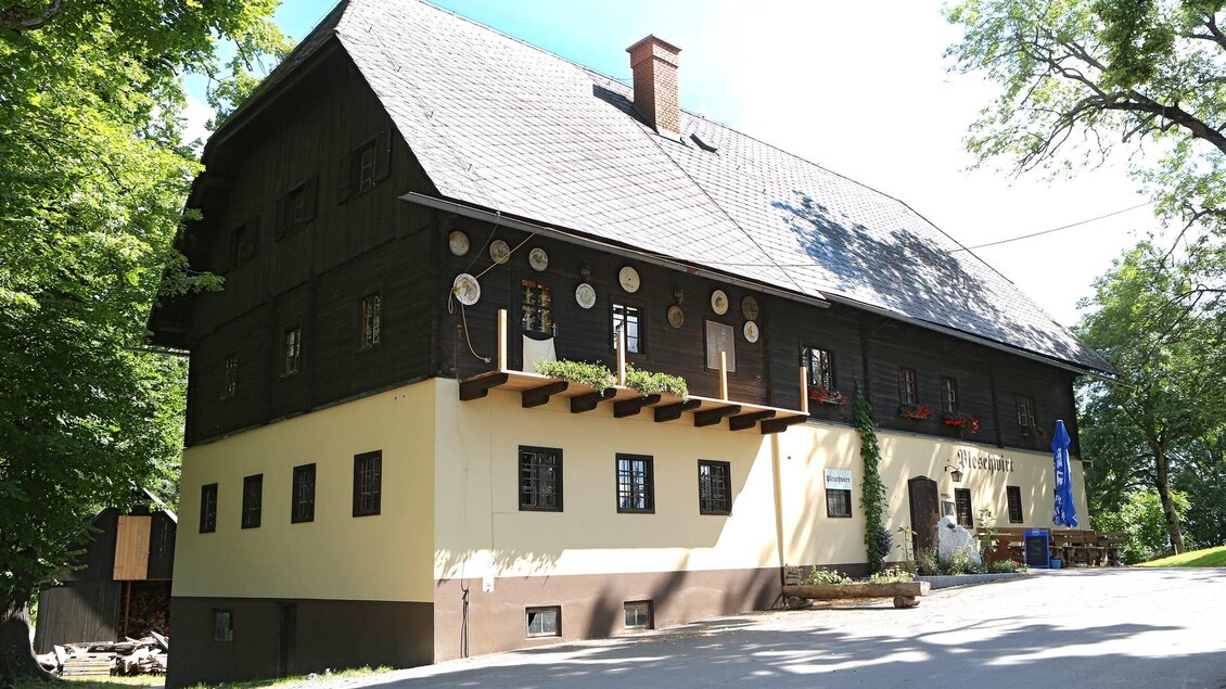 Ein traditionelles, zweigeschossiges Gebäude mit einem schönen, schattigen Außenbereich. Die Fassade ist in zwei Farben gestrichen und wird von Bäumen umgeben. | © TV Region Graz - Donner