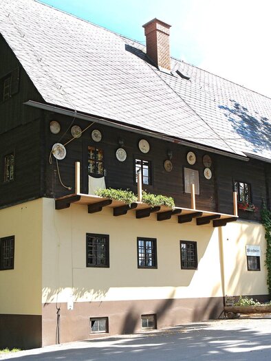 A traditional two-story building with a lovely, shady outdoor area. The facade is painted in two colors and is surrounded by trees. | © TV Region Graz - Donner