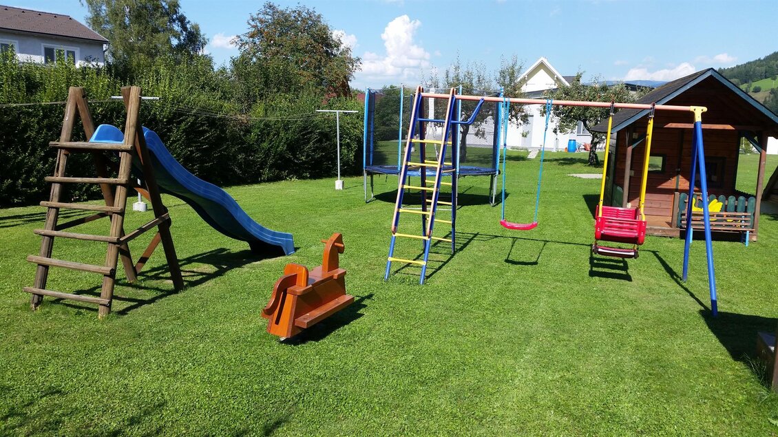 Ein Spielplatz im Freien mit einer Rutsche, Schaukeln und einer kleinen Hütte. Der Garten hat saftig grünes Gras und ist von Bäumen umgeben. | © Gasthaus Rüf-Peterwirt
