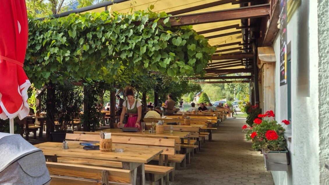 Ein einladendes Restaurant mit vielen Holztischen unter einem begrünten Vordach. Gäste sitzen entspannt und genießen die Atmosphäre. | © Bettina Scheck