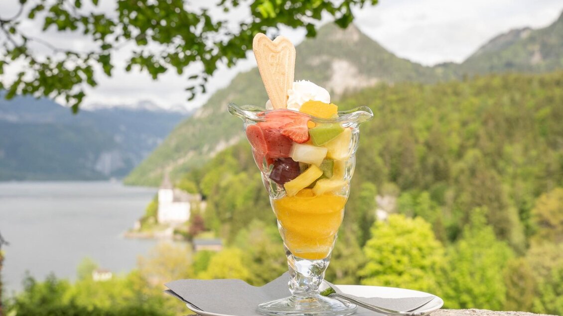 Ein frisches Obstsalat in einem Glas mit verschiedenen bunten Früchten. Im Hintergrund ist eine malerische Landschaft mit einem See und Bergen zu sehen. | © Gasthaus Seeblick
