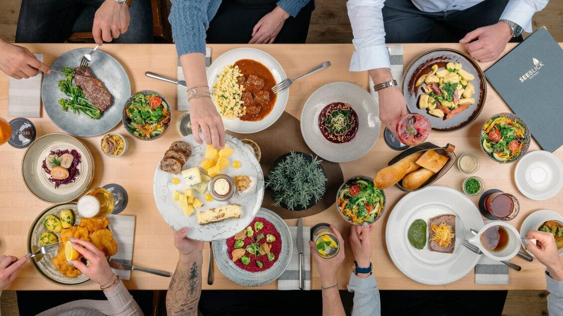 Ein gedeckter Tisch mit verschiedenen Gerichten, darunter Fleisch, Salate und Desserts. Menschen reichen sich das Essen und genießen eine gemeinsame Mahlzeit. | © Gasthaus Seeblick, MONDI Resort
