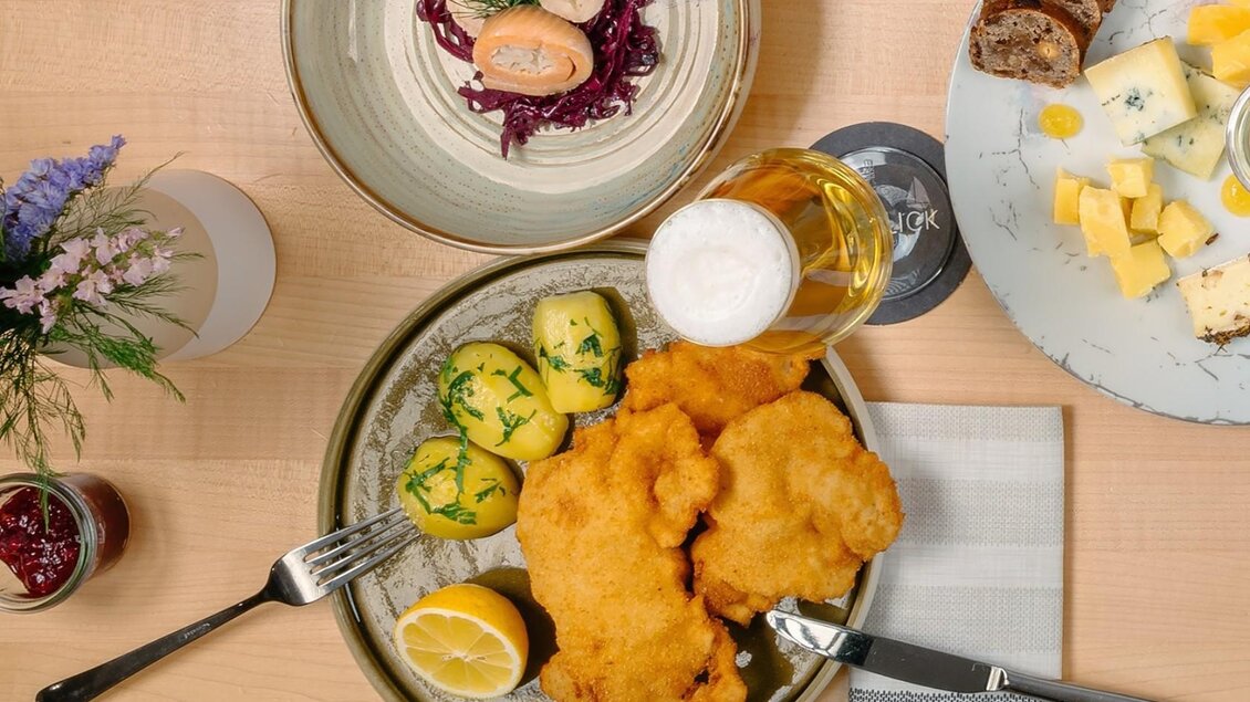 Ein köstliches Gericht mit knusprigem Schnitzel, Kartoffeln und Zitronen auf einem Holztablett. Dazu ein Glas Bier und weitere Beilagen auf dem Tisch. | © Gasthaus Seeblick, MONDI Resort