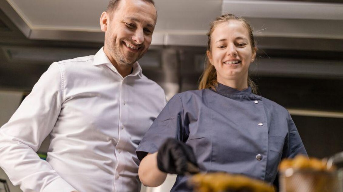 Ein Mann und eine Frau stehen in einer modernen Küche. Die Frau rührt in einem Topf und lächelt, während der Mann zufrieden zuschaut. | © Gasthaus Strobl