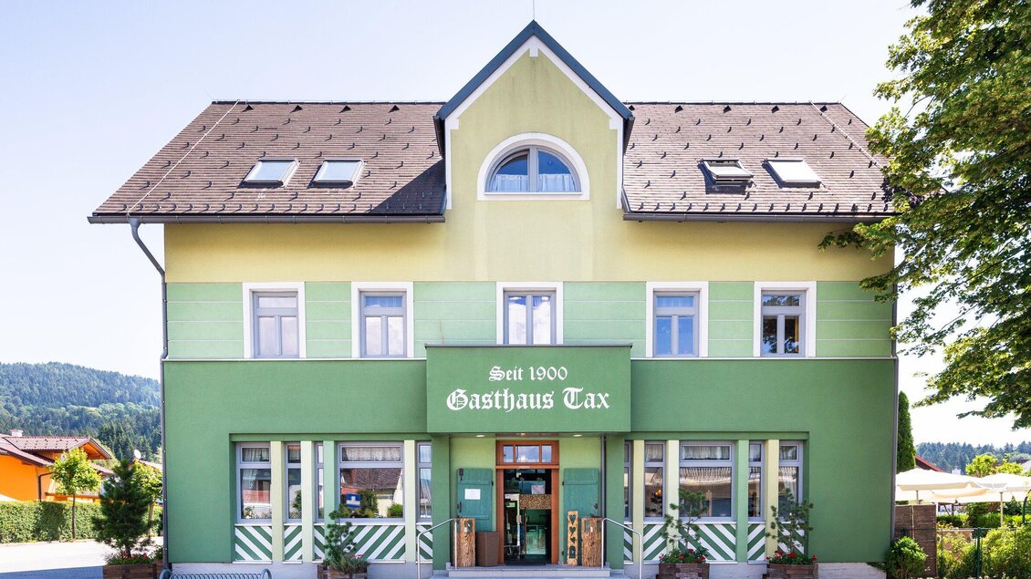 Außenansicht des Gasthauses Tax in Bärnbach, ein traditionelles Gebäude mit grüner Fassade und dem Schriftzug "Gasthaus Tax" im Vordergrund, umgeben von Bäumen und einem sonnigen Himmel. | © Geopho