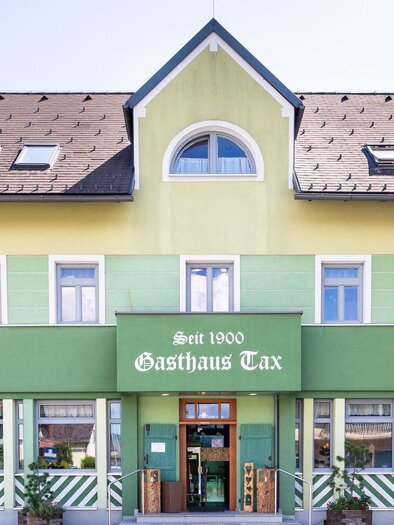 Außenansicht des Gasthauses Tax in Bärnbach, ein traditionelles Gebäude mit grüner Fassade und dem Schriftzug "Gasthaus Tax" im Vordergrund, umgeben von Bäumen und einem sonnigen Himmel. | © Geopho
