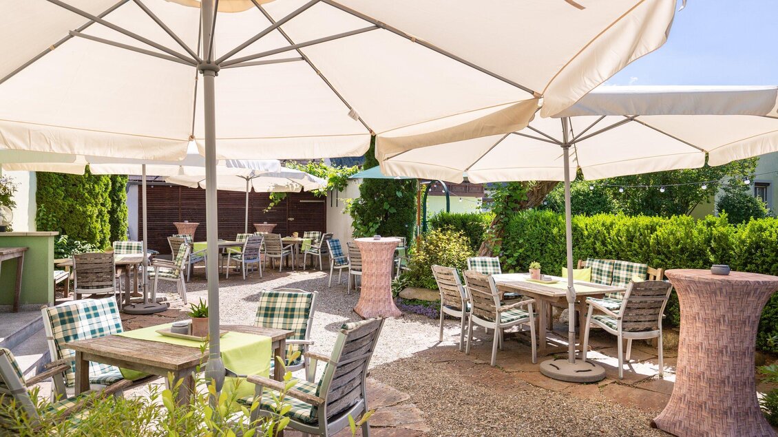 Ruhiger Gastgartenbereich des Gasthaus Tax in Bärnbach mit Tischen unter Sonnenschirmen, umgeben von grünen Pflanzen und einer entspannten Atmosphäre. | © Geopho