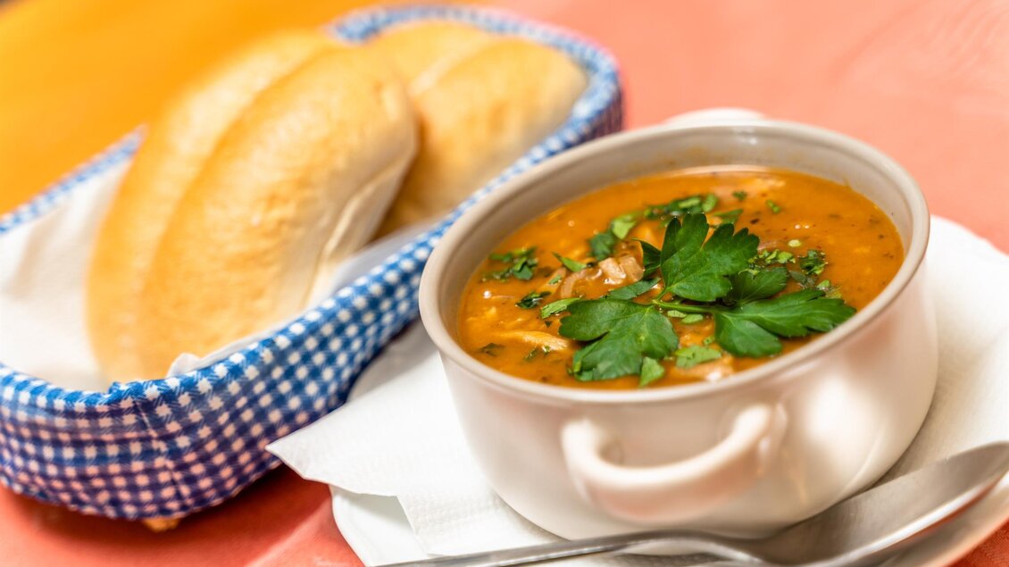 Eine leckere Suppe mit frischer Petersilie in einer weißen Schüssel, begleitet von Brötchen in einem Korb. Die warme Mahlzeit ist auf einem gedeckten Tisch angerichtet. | © Stephan Friesinger