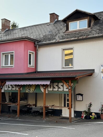 A two-story building with a pink and a white facade. In front of the entrance, there is a covered terrace with seating. | © TV Hochsteiermark
