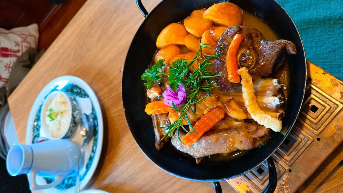 Ein köstliches Gericht in einer gusseisernen Pfanne, das Fleisch mit Pfirsichen und Gemüse enthält. Es ist liebevoll mit Kräutern und einer Blume garniert. | © TV Murau