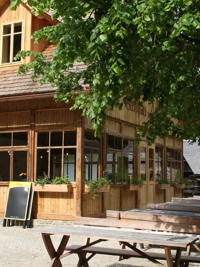 A cozy wooden house with large windows and a green outdoor area. Several wooden tables are set outside, surrounded by trees. | © OeFM Stuebing