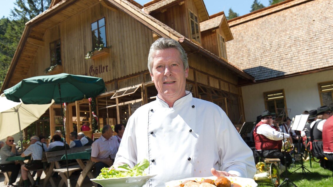 Ein Koch steht mit einem Teller Essen vor einem Holzhaus. Im Hintergrund sind Sitzmöglichkeiten und Gäste zu sehen. | © Region Graz - Harry Schiffer