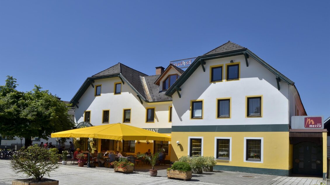 Ein zweistöckiges, gelbes und weißes Gebäude mit einem modernen Design. Vor dem Haus stehen Pflanzen und ein Sonnenschirm bietet Schatten für die Gäste. | © TV Hochsteiermark