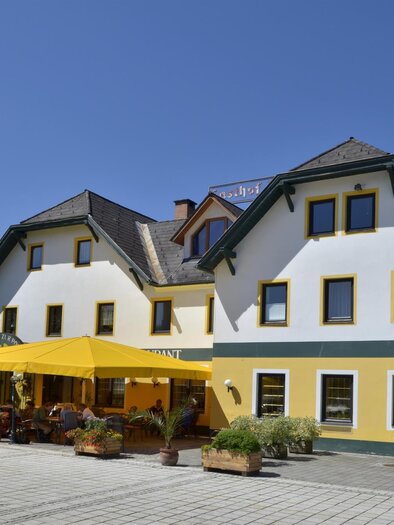 A two-story, yellow and white building with a modern design.  
In front of the house, there are plants and a sunshade provides shade for the guests. | © TV Hochsteiermark