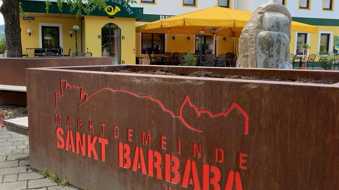 Ein rustikales Schild mit der Aufschrift "Marktgemeinde Sankt Barbara im Mürztal" steht vor einem Gebäude mit gelben Sonnenschirmen. Im Hintergrund sind grüne Bäume und Fenster zu sehen. | © TV Hochsteiermark