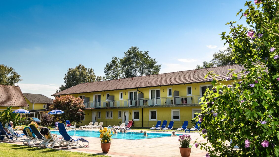 Ein hellgelbes Hotel mit einem Swimmingpool und Sonnenliegen im Garten. Im Hintergrund sind Bäume und blühende Pflanzen zu sehen.