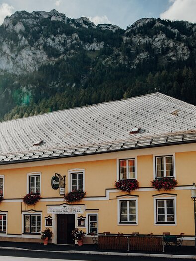 Ein gelbes Gebäude mit Blumenkästen, umgeben von Bergen. Es scheint ein Hotel oder eine Gaststätte zu sein. | © GenussReich