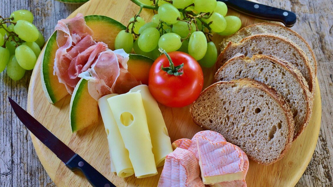 Ein Holzbrett mit verschiedenen Lebensmitteln: Melonenscheiben, Trauben, Schinken, Käse, Tomate und Brotscheiben. Dazu liegen zwei Messer auf dem Brett. | © Symbolfoto Pixabay