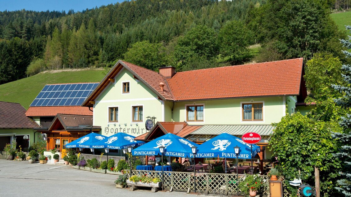 Ein gemütliches Gasthaus in der Natur mit einem roten Dach und grünen Wänden. Vor dem Gebäude stehen Tische mit blauen Sonnenschirmen. | © TV Region Graz - René Vidalli