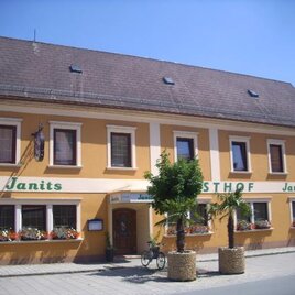 Ein gelb gestrichenes Gebäude mit dem Schriftzug "Janits" und "Gasthof Janits". Vor dem Haus stehen Palmen und Blumen. | © Familie Janits
