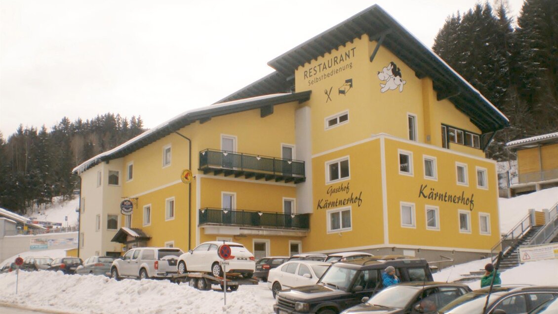 Ein großes gelbes Gebäude mit dem Schriftzug "Restaurant" und "Kärntnerhof" steht vor einer verschneiten Landschaft. Parkplätze sind vor dem Gebäude zu sehen.