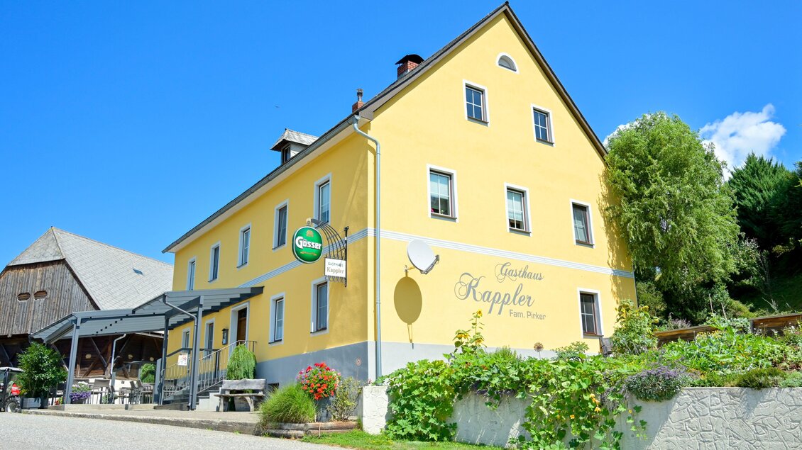 Ein gelbes Gebäude mit mehreren Fenstern und einem Eingang ist zu sehen. Es gibt einen Außenbereich mit Tischen und Stühlen sowie grüne Pflanzen in der Umgebung. | © Gasthof Kappler