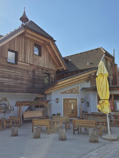 Ein rustikales Gasthaus mit Holzfassade und einer einladenden Terrasse. Im Vordergrund stehen mehrere Holztische und -bänke, umgeben von einer malerischen Landschaft. | © TV Murau