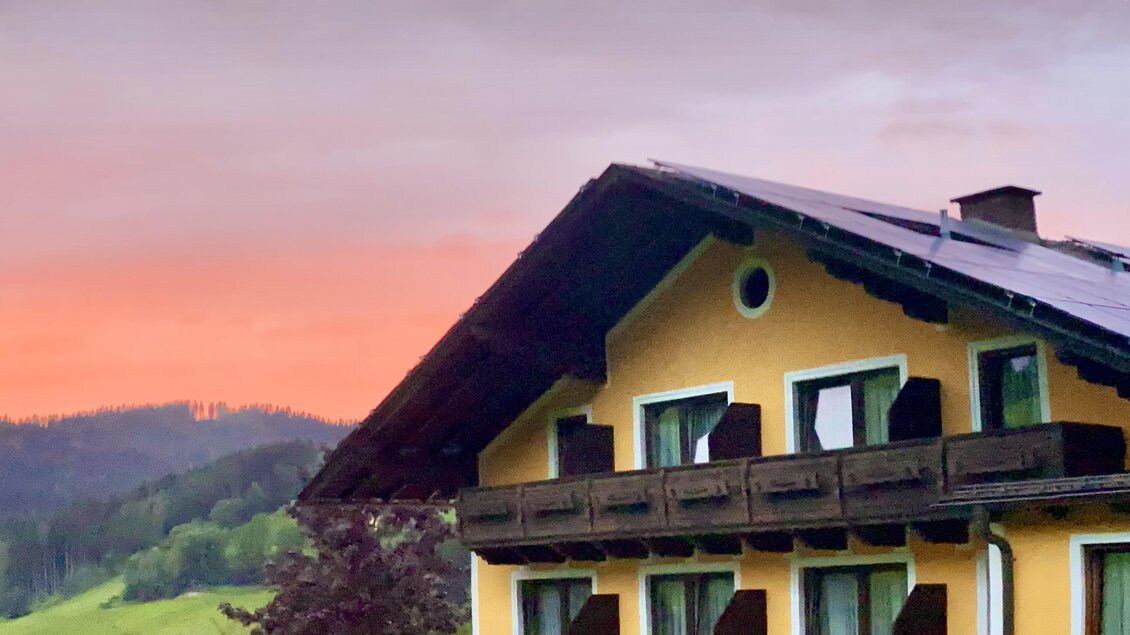Ein charmantes Restaurant in einem gelben Gebäude mit Blick auf die grünen Hügel. Im Hintergrund erstrahlt der Himmel in warmen Farben bei Sonnenuntergang. | © Gasthof Landhotel Hubmann
