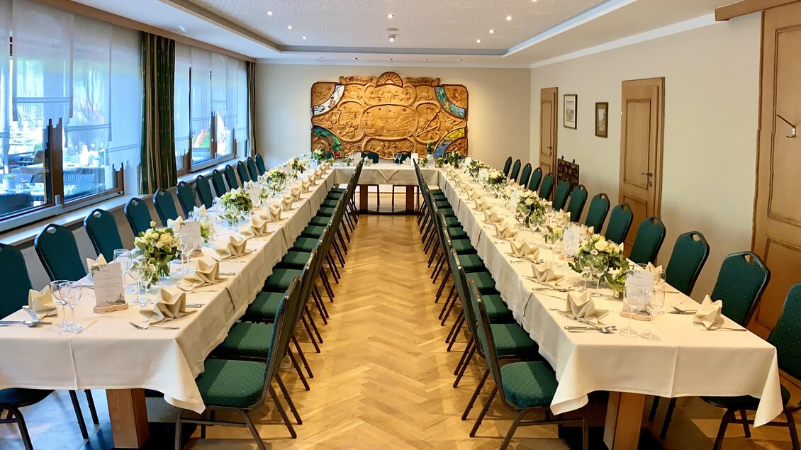 Ein eleganter Speisesaal mit langen Tischen, dekoriert mit Blumen und weißer Tischwäsche. Im Hintergrund ist ein kunstvolles Wandrelief zu sehen. | © Gasthof Landhotel Hubmann