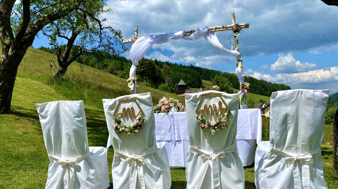 Eine romantische Hochzeitsdekoration im Freien mit weißen Stühlen und einem geschmückten Altar. Die Landschaft ist grün und der Himmel strahlt blau mit einigen Wolken. | © Gasthof Landhotel Hubmann
