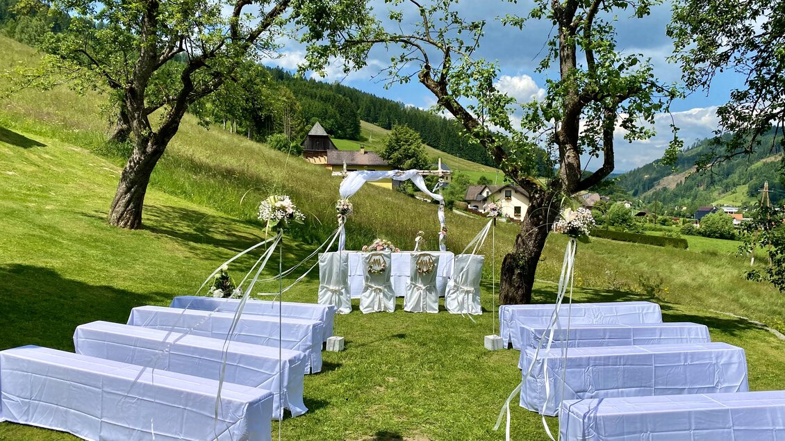 Eine romantische Hochzeit im Freien mit weißen Bänken und einer festlichen Dekoration. Im Hintergrund sind grüne Wiesen und Berge zu sehen. | © Gasthof Landhotel Hubmann